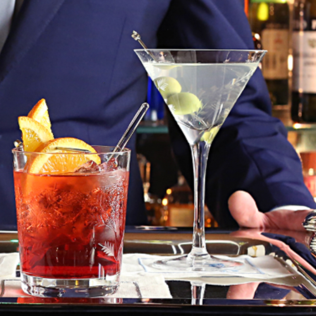 A person in a dark suit holds a tray with a Negroni and a martini garnished with green olives, served in the William Yeoward Vesper Martini Glass, set before a bar.