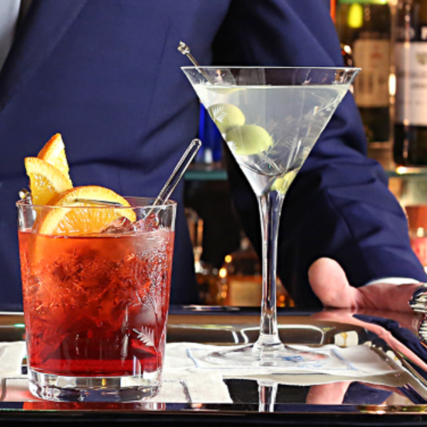 A person in a dark suit holds a tray with a Negroni and a martini garnished with green olives, served in the William Yeoward Vesper Martini Glass, set before a bar.