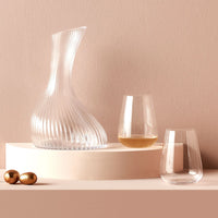 The Stem Zero Vertigo Water Glass Set of 2 by Nude Glassware is displayed on a curved cream platform with gold decorative spheres nearby, one glass containing white wine, all set against a beige background.