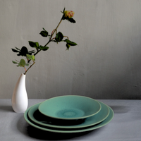 The Vuelta Nori plates by Jars - Rosenthal, shown stacked in turquoise on a gray surface beside a white vase with a single rose, are modern serving pieces ideal for everyday use.