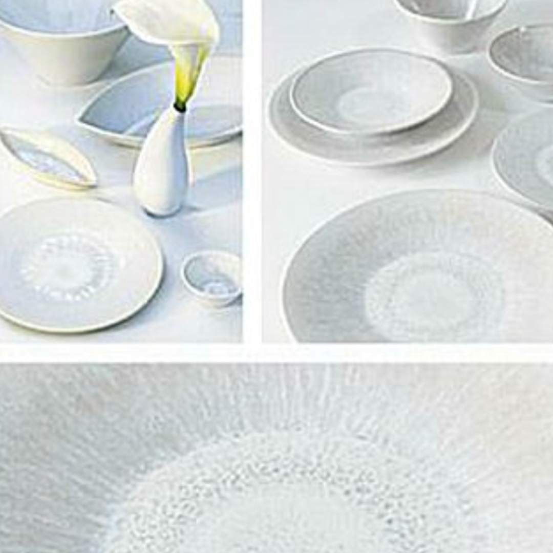 The Vuelta White Pearl collection by Jars - Rosenthal features white ceramic dishes and bowls with subtle blue patterns, plus an atoll-glazed dinner plate, all displayed with a white vase holding a single calla lily.