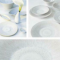 The Vuelta White Pearl collection by Jars - Rosenthal features white ceramic dishes and bowls with subtle blue patterns, plus an atoll-glazed dinner plate, all displayed with a white vase holding a single calla lily.