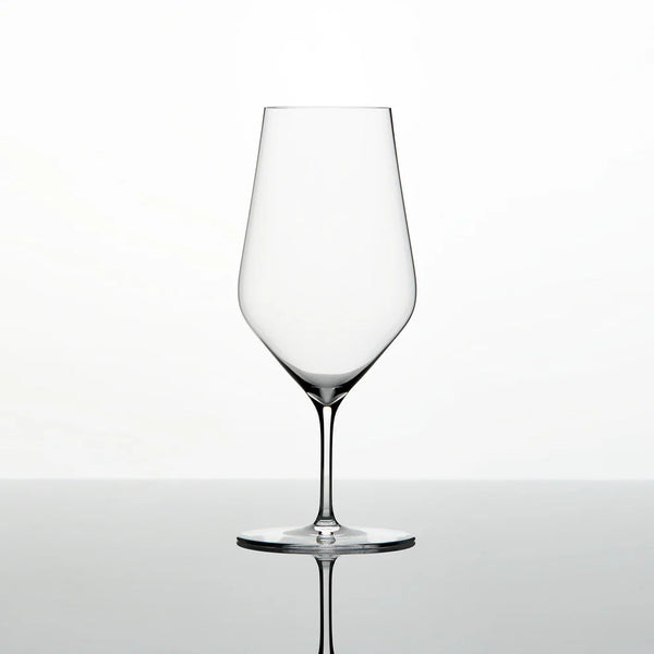 A single Zalto Water Glass by Grand Cru Selections / Zalto, featuring a thin stem and wide bowl, stands empty on a reflective surface against a plain white background.