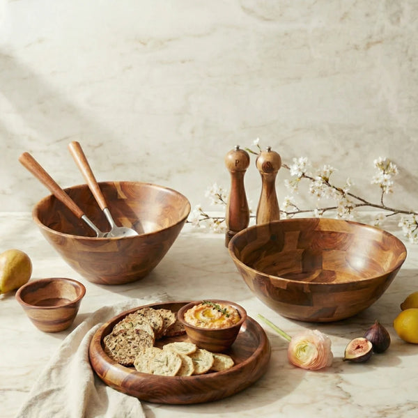 The Be Home Rosewood Snack Bowl is crafted from sustainably sourced wood, highlighting its natural grain. It’s featured on a marble surface with flowers, surrounded by serving accessories like salad utensils and grinders.