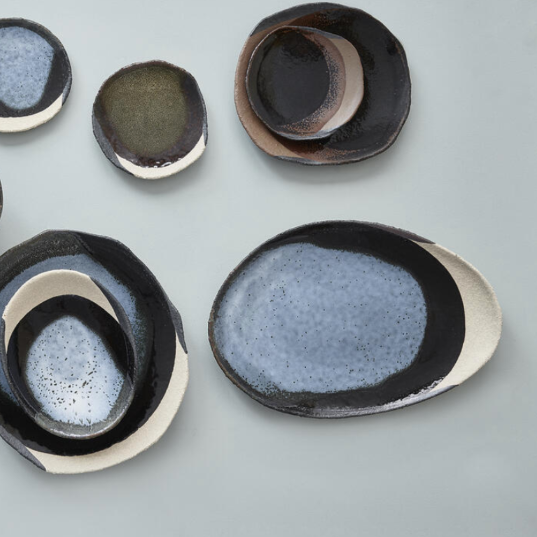 Wabi Kemuri by Jars: hand-crafted ceramic plates and bowls in organic shapes, featuring blue, black, white, and brown tones. Artistically arranged on light grey, inspired by Japanese Wabi Sabi aesthetics.