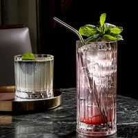 Two cocktails on a dark marble table: a clear drink with mint in a NUDE Wayne DOF Glass from the Nude Glassware Wayne DOF Glass Set of 4, and a tall pink drink with ice, mint, and metal straw in a lead-free crystal highball glass.