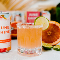 A glass of grapefruit paloma with a salted rim and lime garnish sits in front of a can labeled June Shine, served in a Wayne DOF Glass from Nude Glassware, surrounded by sliced grapefruit and lime.