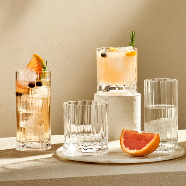 The Wayne Highball Glass Set of 4 by Nude Glassware is displayed on a textured surface, featuring two glasses with citrus and herb cocktails, one with water and ice, and one empty, accented by a grapefruit wedge in soft natural light.