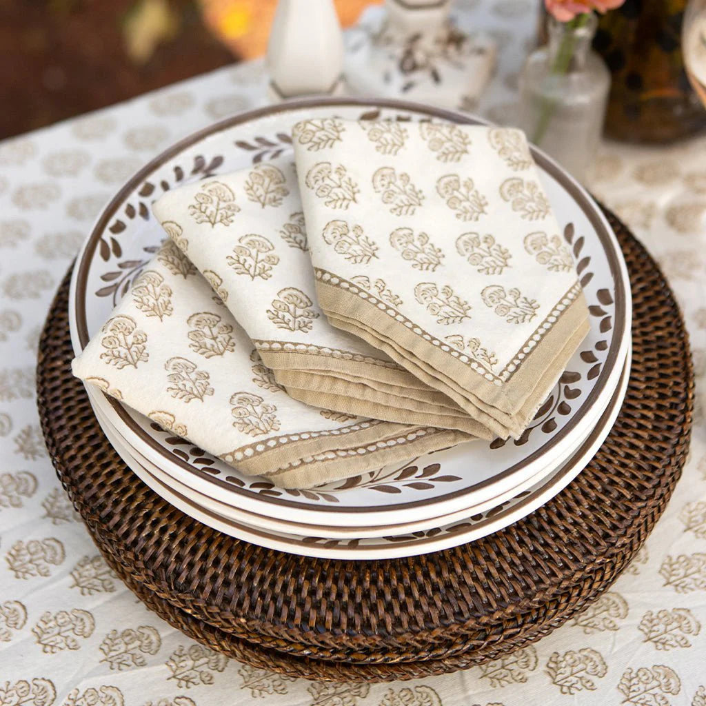 A set of Pomegranate's Anika Napkin Set of 4 in beige, featuring block print florals, is neatly arranged on brown-accented white plates atop a round woven placemat and a patterned tablecloth.
