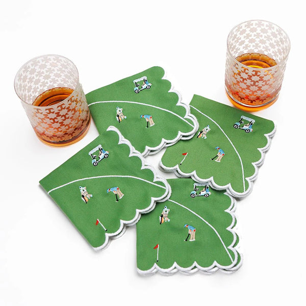 Two glasses rest beside the Day At The Golf Course Cocktail Napkin Set of 4 by Pomegranate—green, scalloped-edge napkins featuring golfers, golf carts, and flags—to add playful charm to your tableware on a crisp white background.