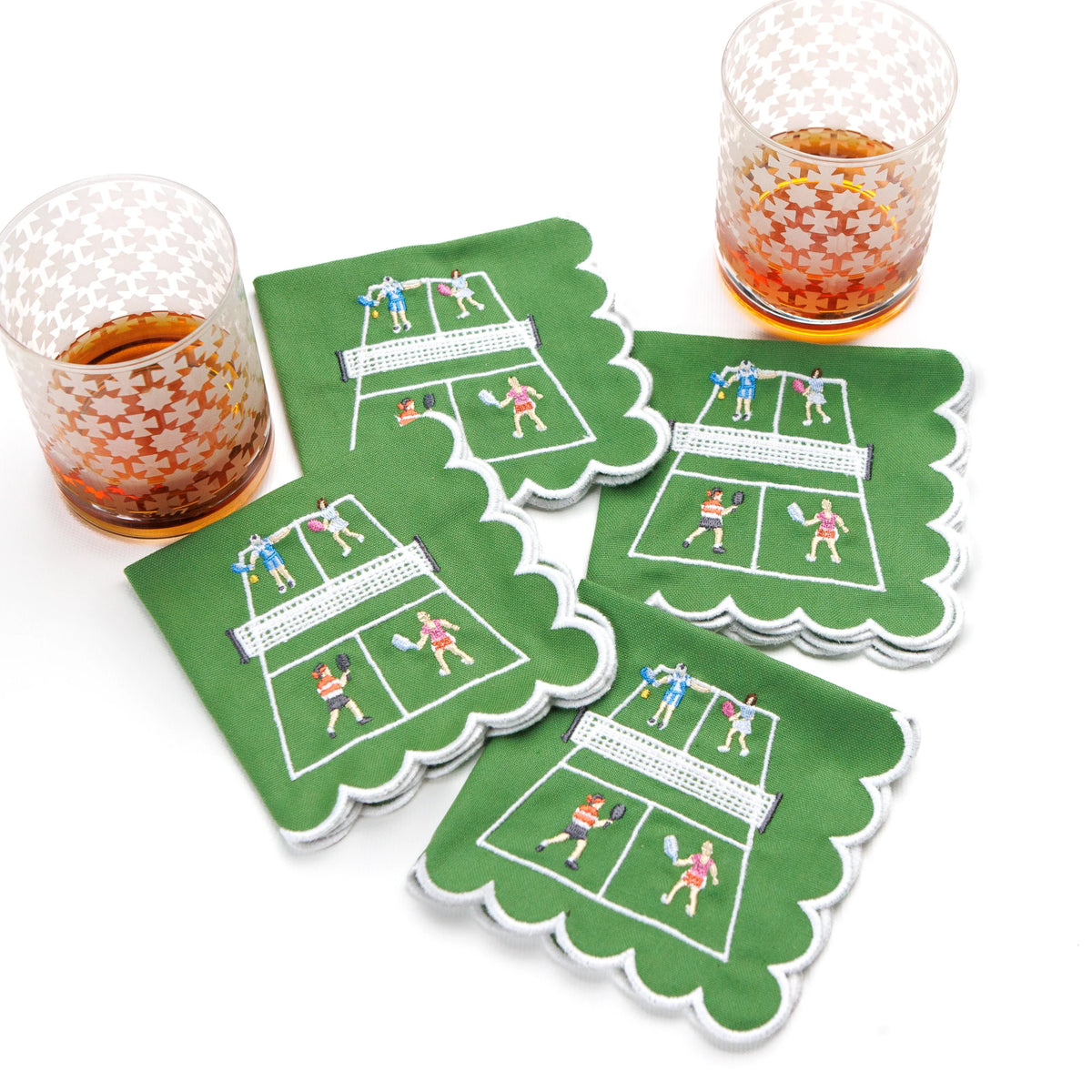 The Day At The Courts Cocktail Napkin Set of 4 by Pomegranate features green cotton napkins with scalloped edges and playful tennis motifs, shown beside two patterned glasses of amber drinks on a white surface.