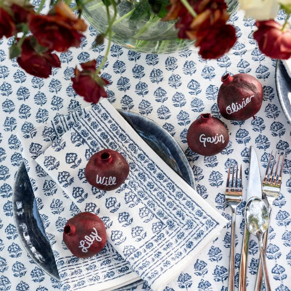 A table setting features Pomegranate’s Elise Napkin Set of 4 - Blue, layered blue tableware, silverware, red pomegranate place cards for Will, Lily, Gavin, and Olivia, with red flowers hanging above.