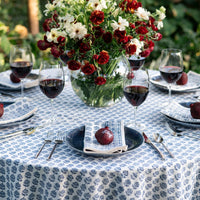 A round table set for four features wine glasses, plates, silverware, and red apples atop Pomegranate's Elise Napkin Set of 4 – Blue. A vase of red and white flowers sits on a blue and white floral cloth, enhancing the handcrafted decor.