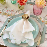 A pastel table setting with a blue plate, Bodrum Firenze Napkin Set of 4 - Ice Blue scalloped napkin held by a gold floral ring, vintage-style cutlery, pink glass, topped off with a floral runner and vases of colorful blooms.