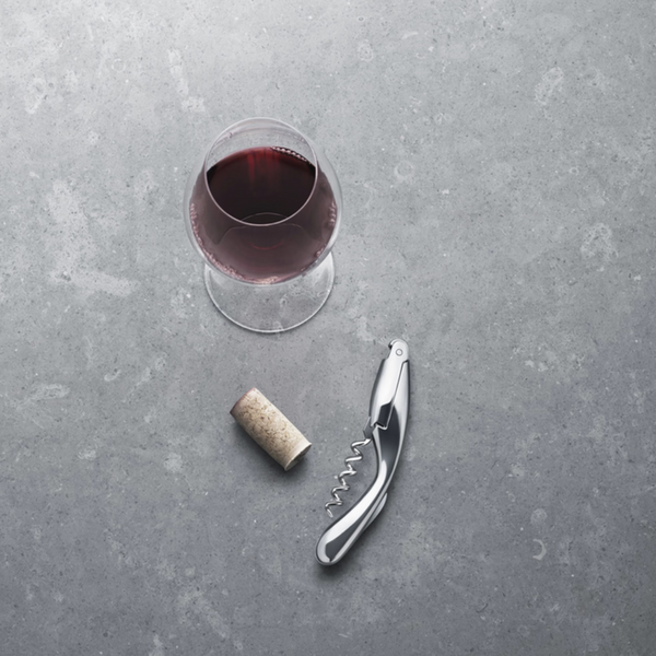A glass of red wine sits on a gray surface next to a cork and the elegant Georg Jensen Wine and Bar Corkscrew.
