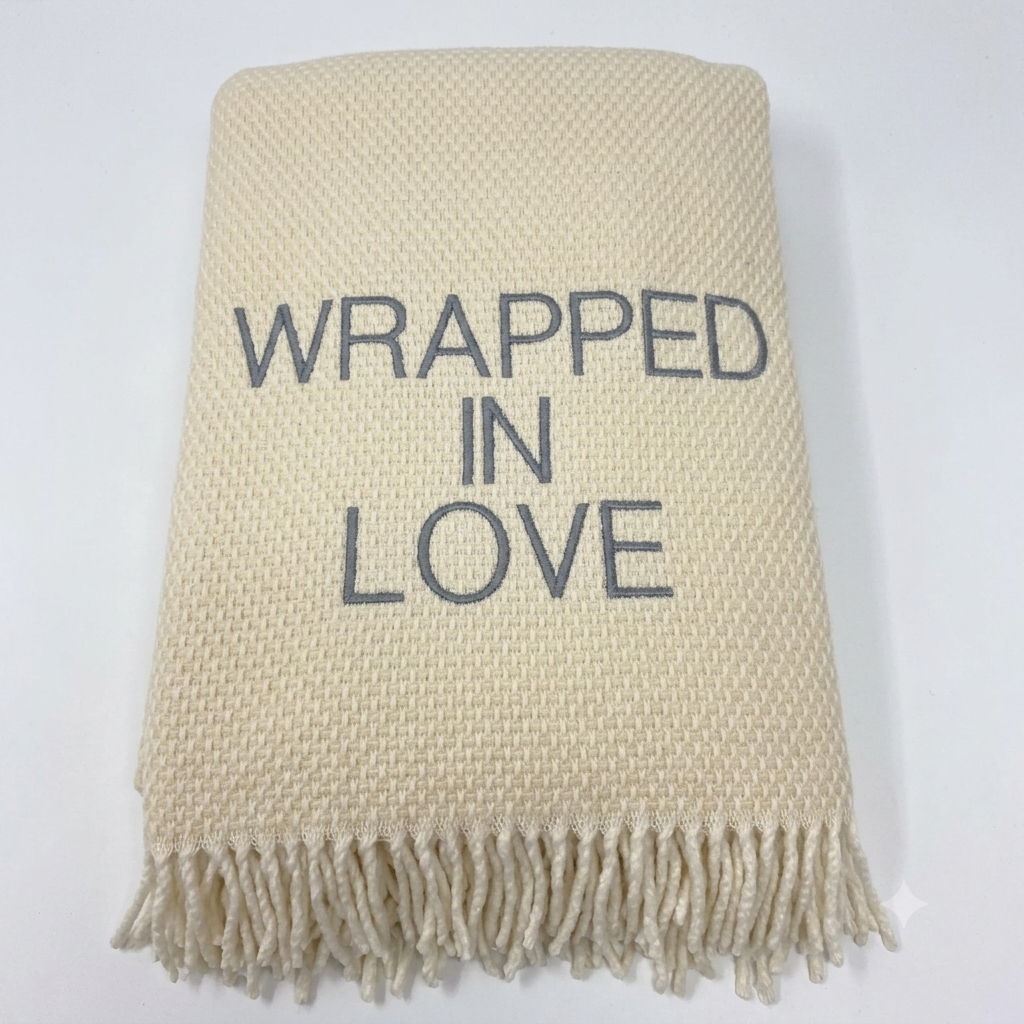 The Wrapped in Love Throw by A Soft Idea is a super soft, cream-colored woven blanket with fringed edges and "WRAPPED IN LOVE" embroidered in gray—an ideal holiday gift.
