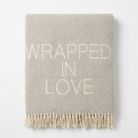 The A Soft Idea Wrapped in Love Throw is a beige woven blanket with fringe and "WRAPPED IN LOVE" in bold white letters—perfect as a cozy, thoughtful holiday gift.