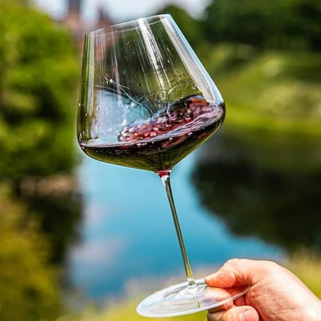 A hand holds the Zalto Burgundy Glass by Grand Cru Selections / Zalto, swirling red wine as trees and blue sky reflect in the background, showcasing the elegance of this lead-free wine glass.