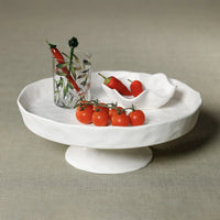 The Zodax Alto Ceramic Pedestal Bowl displays cherry tomatoes and a small dish of red chili peppers. Next to it, a glass with green leaf design contains more chilies, all set against a simple neutral background.