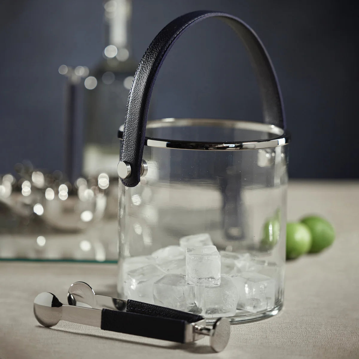 The Zodax Laguna Ice Bucket features a glass design with a black handle, filled with ice cubes. Metal tongs with black grips rest in front, while limes, a glass bottle, and barware accent the scene. Ideal as a wine or champagne chiller.