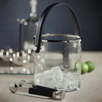 The Zodax Laguna Ice Bucket features a glass design with a black handle, filled with ice cubes. Metal tongs with black grips rest in front, while limes, a glass bottle, and barware accent the scene. Ideal as a wine or champagne chiller.
