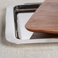 A close-up of the Braxton Wood Serving Tray by Zodax, a rectangular wooden board resting atop a shiny metal tray, both placed on a textured light-colored fabric surface.