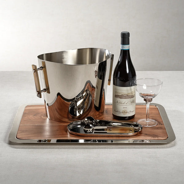 A silver ice bucket, a wine bottle, a wine glass, and a corkscrew are elegantly arranged on the Zodax Braxton Wood Serving Tray, which features a silver border and rests atop a light-colored tablecloth.