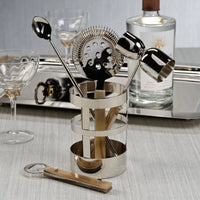 The Zodax Belmont 5 Piece Bar Tool Set features a jigger, strainer, bar spoon, and bottle opener in a shiny holder—ideal for upgrading your home bar décor. Glasses and a bottle complete the stylish scene.