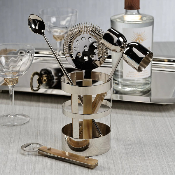 The Zodax Belmont 5 Piece Bar Tool Set features a jigger, strainer, bar spoon, and bottle opener in a shiny holder—ideal for upgrading your home bar décor. Glasses and a bottle complete the stylish scene.