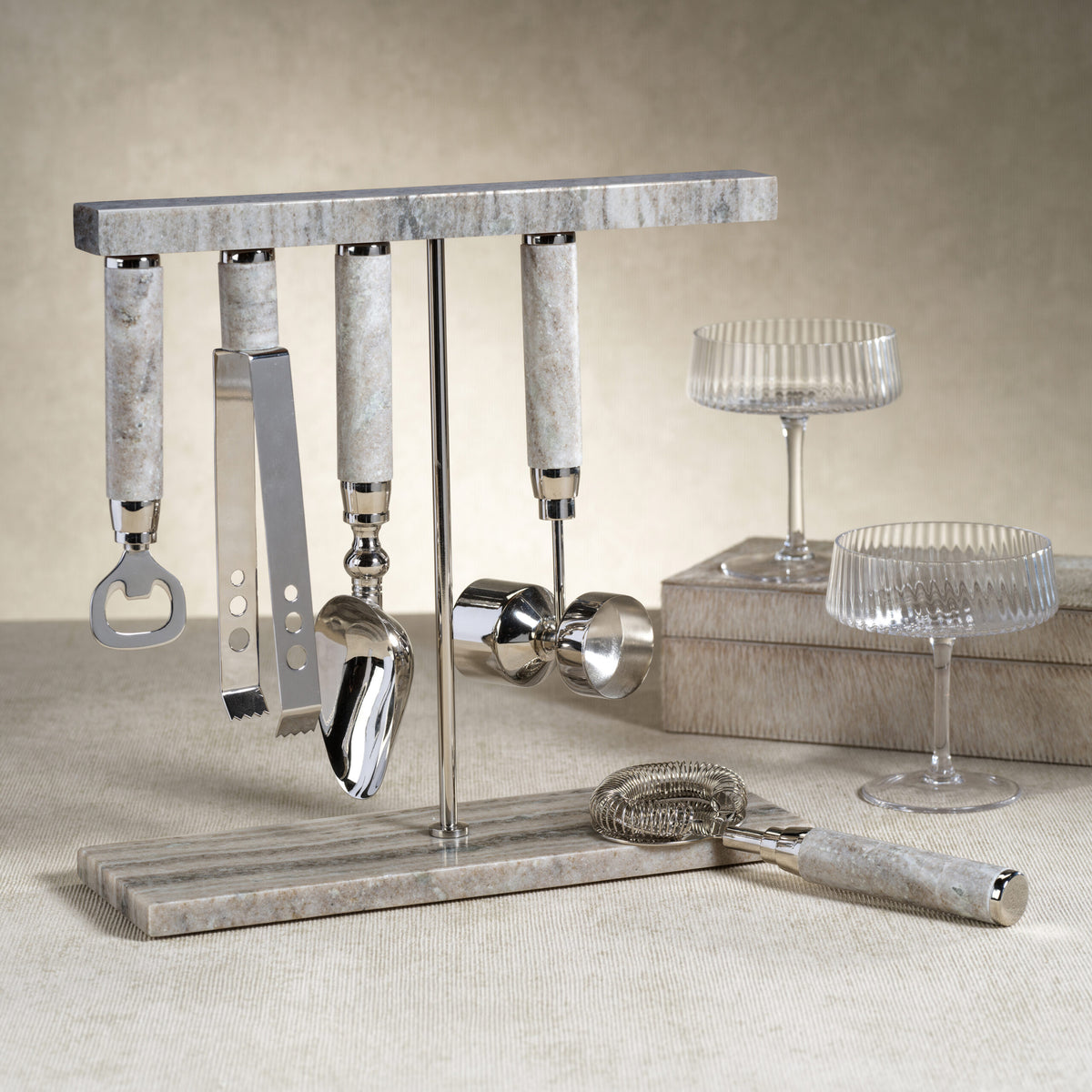 The Zodax Grey Marble Bar Tool 5 Piece Set includes stainless steel tools—a bottle opener, ice tongs, scoop, jigger, and strainer—on a matching stand, with two ribbed cocktail glasses on marble coasters in the background.