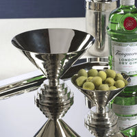 A silver martini glass, the Zodax Malcolm Condiment Bowl in polished nickel filled with green olives, a bottle of Tanqueray gin, and a cocktail shaker are arranged on a reflective tray in striking Art Deco style.