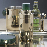 The Zodax Spector Cocktail Shaker in gold and green is displayed on a tray with matching gold barware, a bottle of Chartreuse, green coasters, and a gold ice bucket featuring green handles.