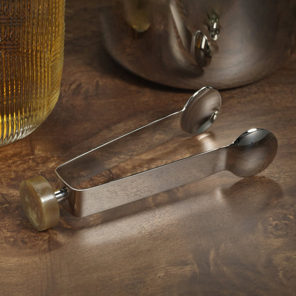 The Zodax Spector Ice Tongs - Nickel & Amber feature polished nickel with an amber marbled handle, shown resting on wood near a glass of yellow liquid and metal pot—a refined bar accessory.