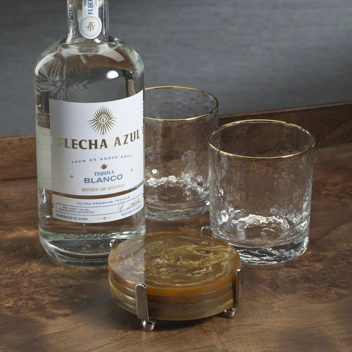 A bottle of Flecha Azul tequila sits on a wooden surface next to two gold-rimmed glasses and a Zodax Spector Round Coaster Set - Amber displayed on a modern host’s polished nickel stand.