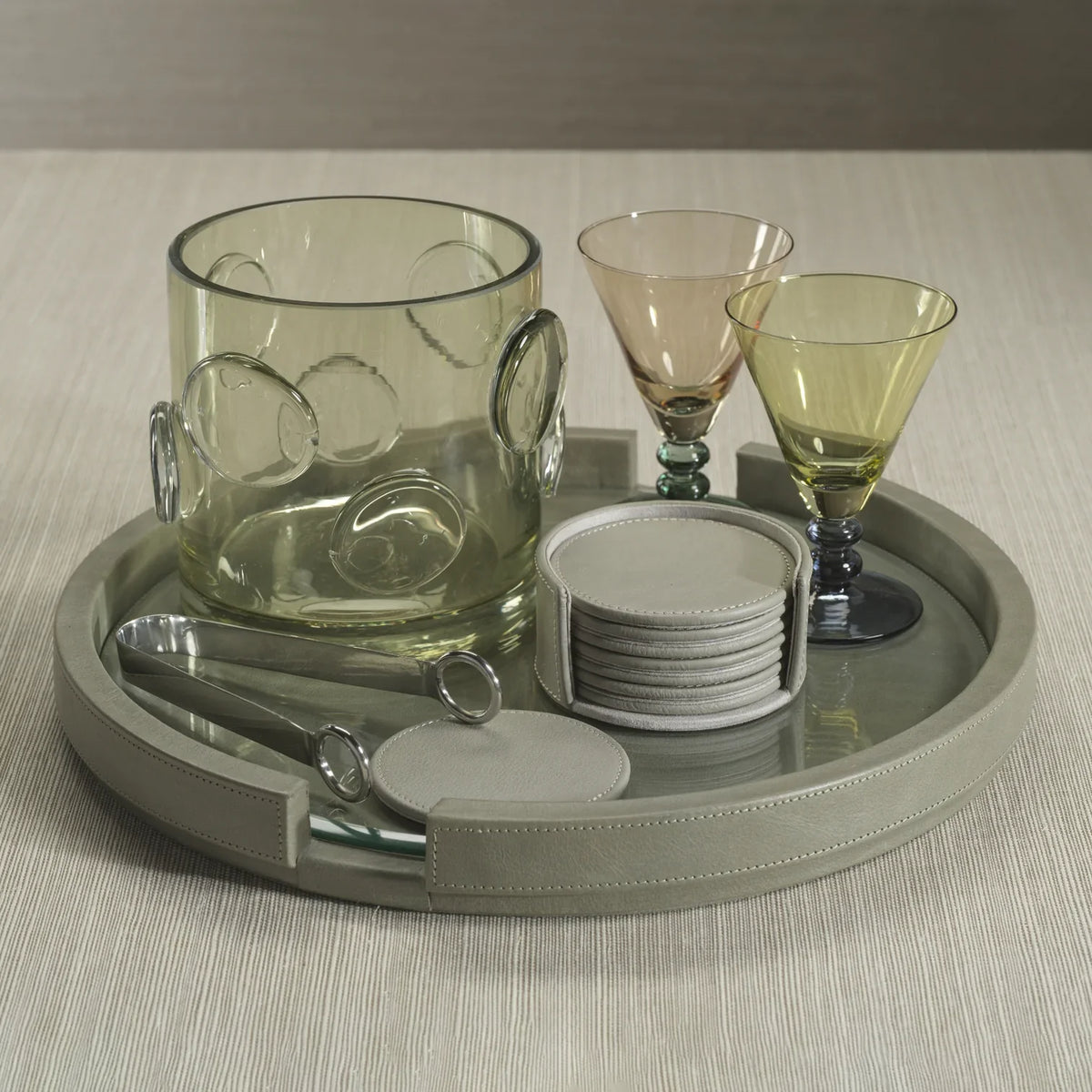 A round tray displays a textured glass ice bucket, two stemmed glasses, tongs, a cocktail stirrer, and the Zodax Capri Coaster Set of 8 in grey, all arranged neatly on a light textured surface.
