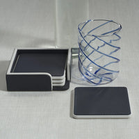 The Zodax Napoli Leather Coaster Set of 4 in navy and white sits in its holder beside a clear glass with blue spiral stripes on a light surface, with one coaster from the set placed outside the holder.