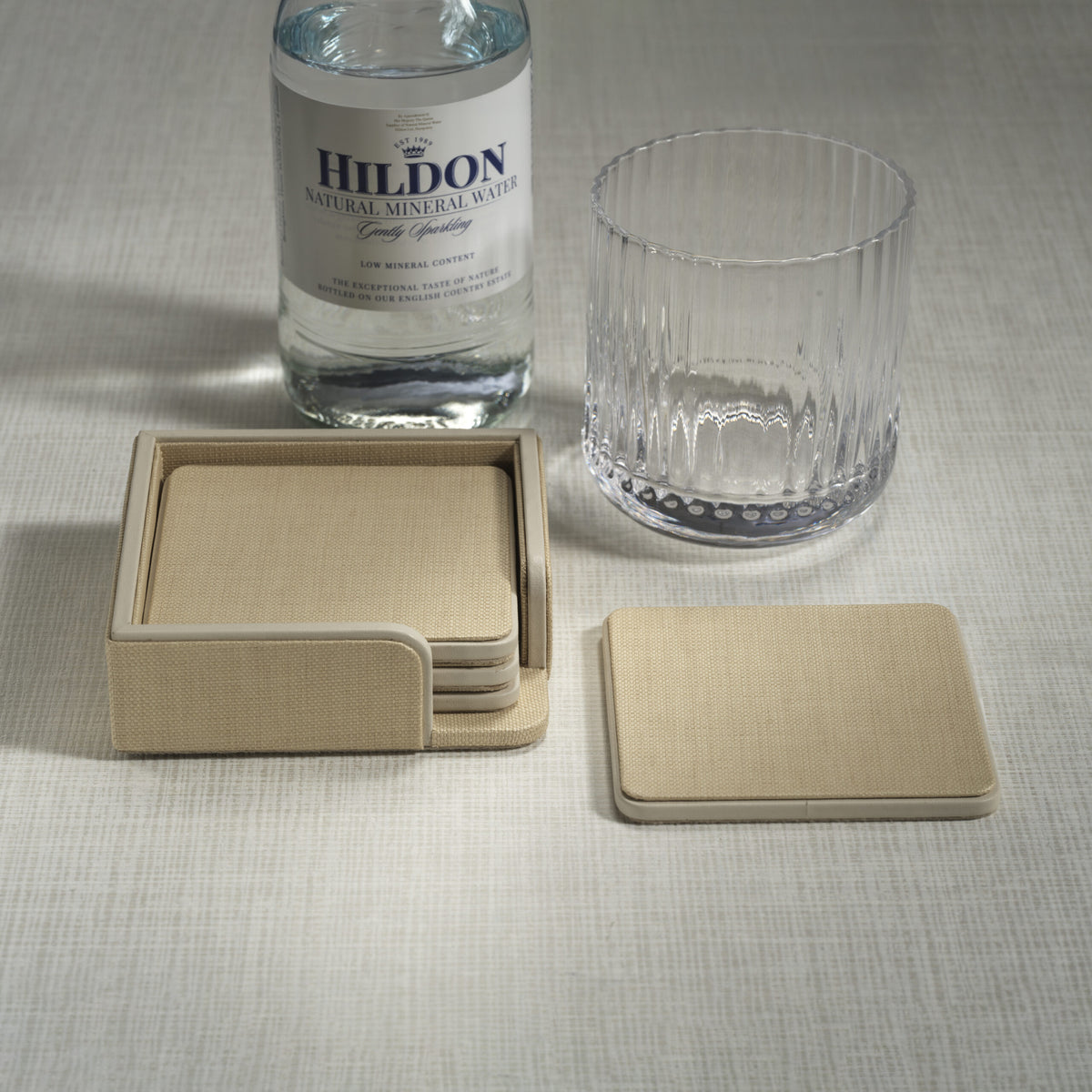 A bottle of Hildon mineral water, an empty faceted glass, and a Zodax Napoli Leather Coaster Set of 4 in Raffia & Cream are arranged on a light textured surface, with one coaster placed outside the matching holder.