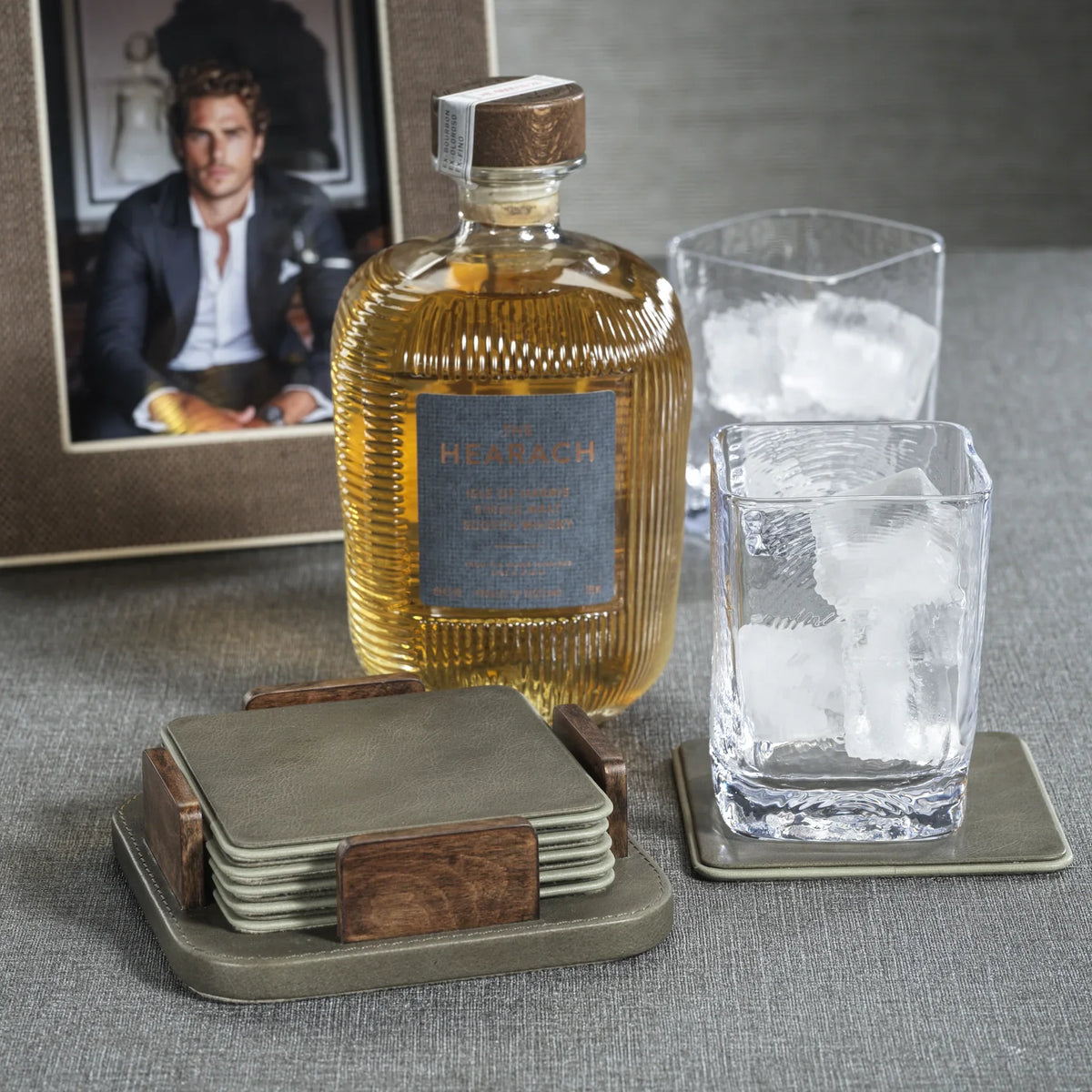 A bottle of whiskey, two glasses with ice, the Zodax Leather & Veneer Coaster Set - Cedar Green, and a framed photo of a man are arranged on a gray surface.
