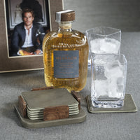 A bottle of whiskey, two glasses with ice, the Zodax Leather & Veneer Coaster Set - Cedar Green, and a framed photo of a man are arranged on a gray surface.