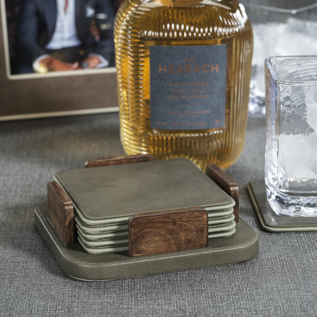 A Zodax Leather & Veneer Coaster Set - Cedar Green sits on the table beside a whiskey bottle, an empty glass with ice, and a framed photo in the background.