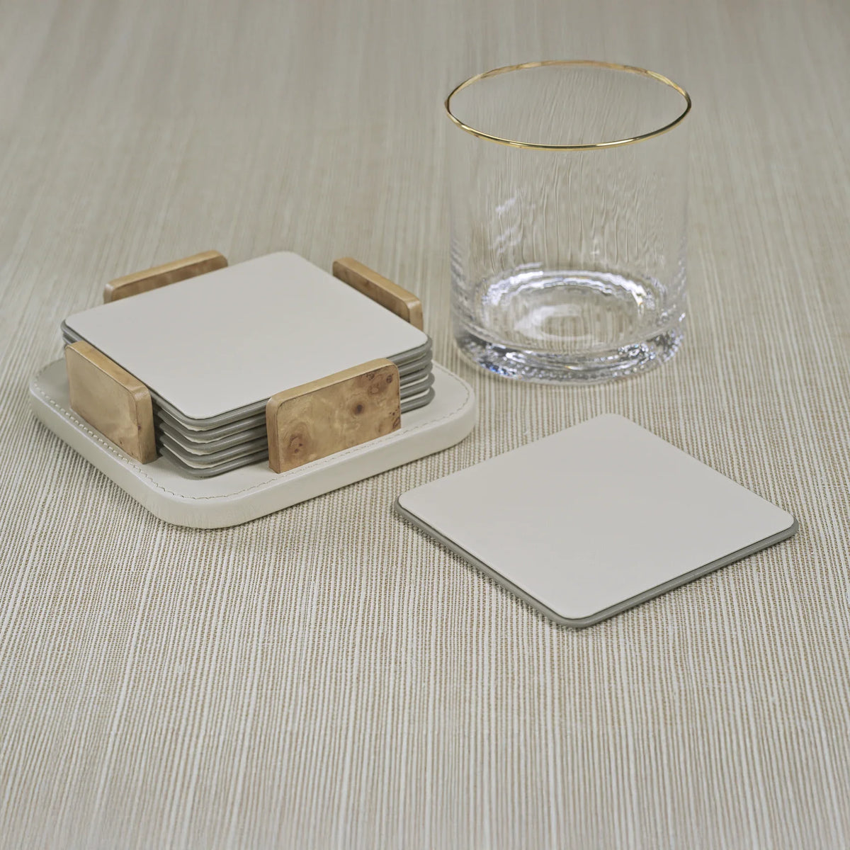 The Zodax Leather & Veneer Coaster Set in Ivory, paired with wooden holders, is displayed beside a clear glass with a gold rim and a single matching coaster on a light textured surface.