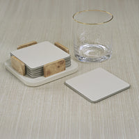 The Zodax Leather & Veneer Coaster Set in Ivory, paired with wooden holders, is displayed beside a clear glass with a gold rim and a single matching coaster on a light textured surface.