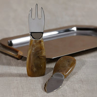 The Borghese Cheese Set by Zodax features two serving utensils with marbled brown handles—one fork and one spreader—resting before a shiny metal tray, ideal for your charcuterie board on a beige fabric surface.