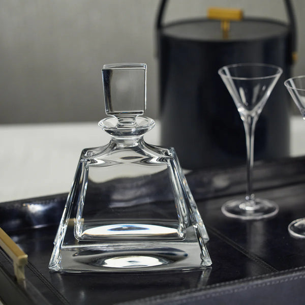 The Zodax Noelle Crystal Decanter, featuring a square silhouette and elegant stopper, rests on a black tray alongside two empty martini glasses and a black ice bucket with a gold handle—creating a sophisticated addition to any home bar.