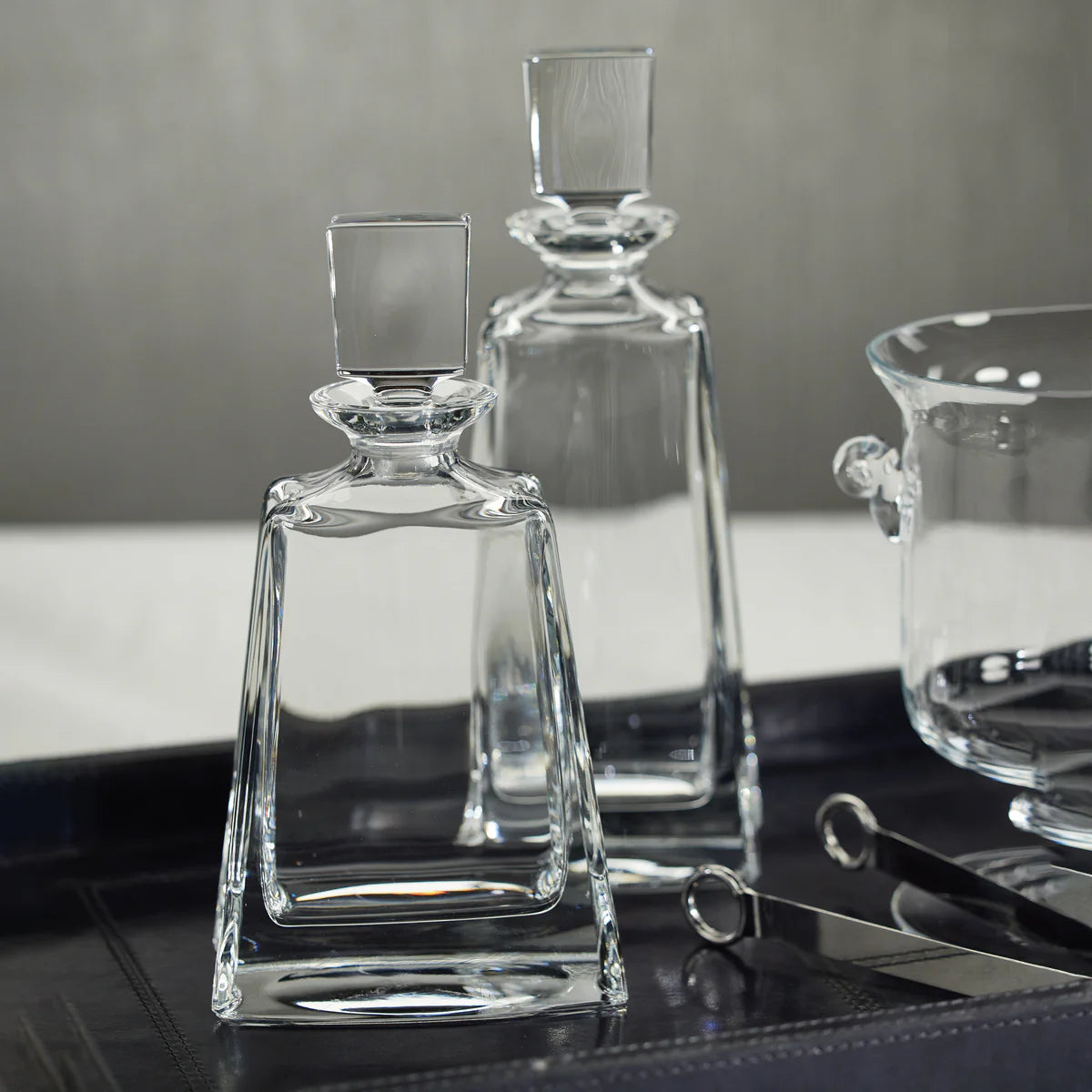 Two Kelsey Crystal Decanters by Zodax, each featuring a clear glass design with square stoppers, sit on a dark tray beside a glass bowl and metal tongs against a neutral backdrop. An elegant addition to any home bar or barware collection, these Zodax crystal decanters elevate your entertaining style.