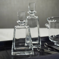 Two Kelsey Crystal Decanters by Zodax, each featuring a clear glass design with square stoppers, sit on a dark tray beside a glass bowl and metal tongs against a neutral backdrop. An elegant addition to any home bar or barware collection, these Zodax crystal decanters elevate your entertaining style.