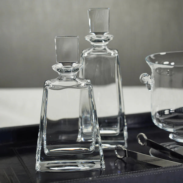 Two Kelsey Crystal Decanters by Zodax, each featuring a clear glass design with square stoppers, sit on a dark tray beside a glass bowl and metal tongs against a neutral backdrop. An elegant addition to any home bar or barware collection, these Zodax crystal decanters elevate your entertaining style.