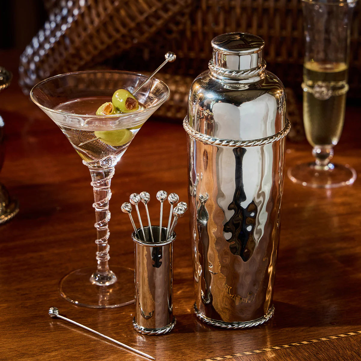 The Juliska Graham Cocktail Shaker is elegantly arranged on a polished wooden surface beside a martini garnished with green olives, silver stirring sticks, and a glass of champagne.