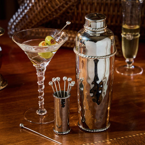 The Juliska Graham Cocktail Shaker is elegantly arranged on a polished wooden surface beside a martini garnished with green olives, silver stirring sticks, and a glass of champagne.