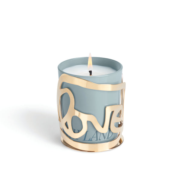 The Love Candle - Upland by Amber Sakai features a lit white candle in a light blue holder, adorned with a decorative gold metal sleeve that spells “LOVE” in elegant cursive letters.